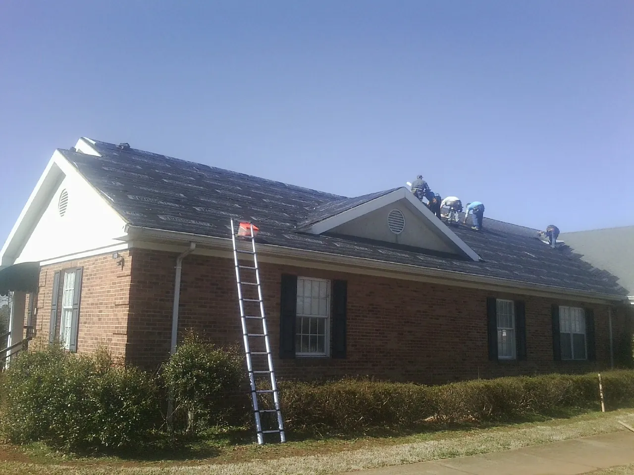 Crew tearing shingles off a brick ranch roof with a ladder set — Metal Roof Repair work by our crew in Baker